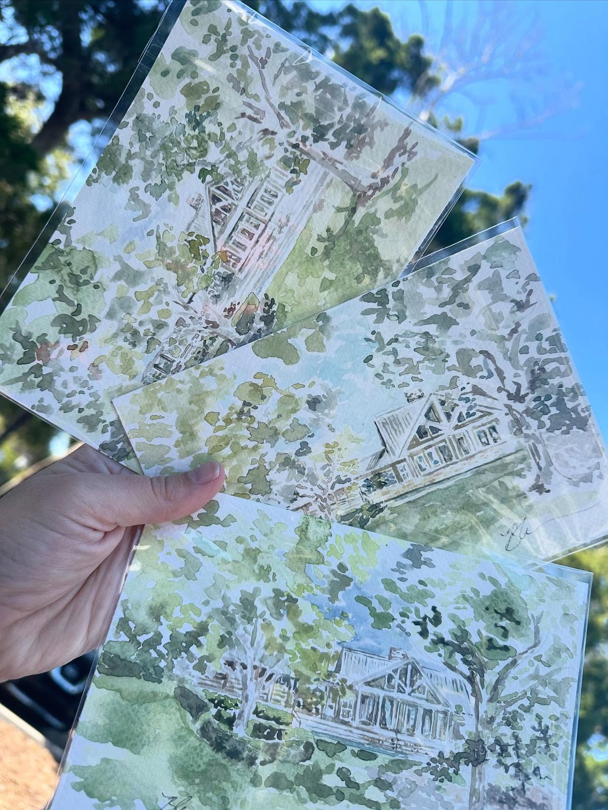 Three finished watercolor house portraits held up against a blue sky