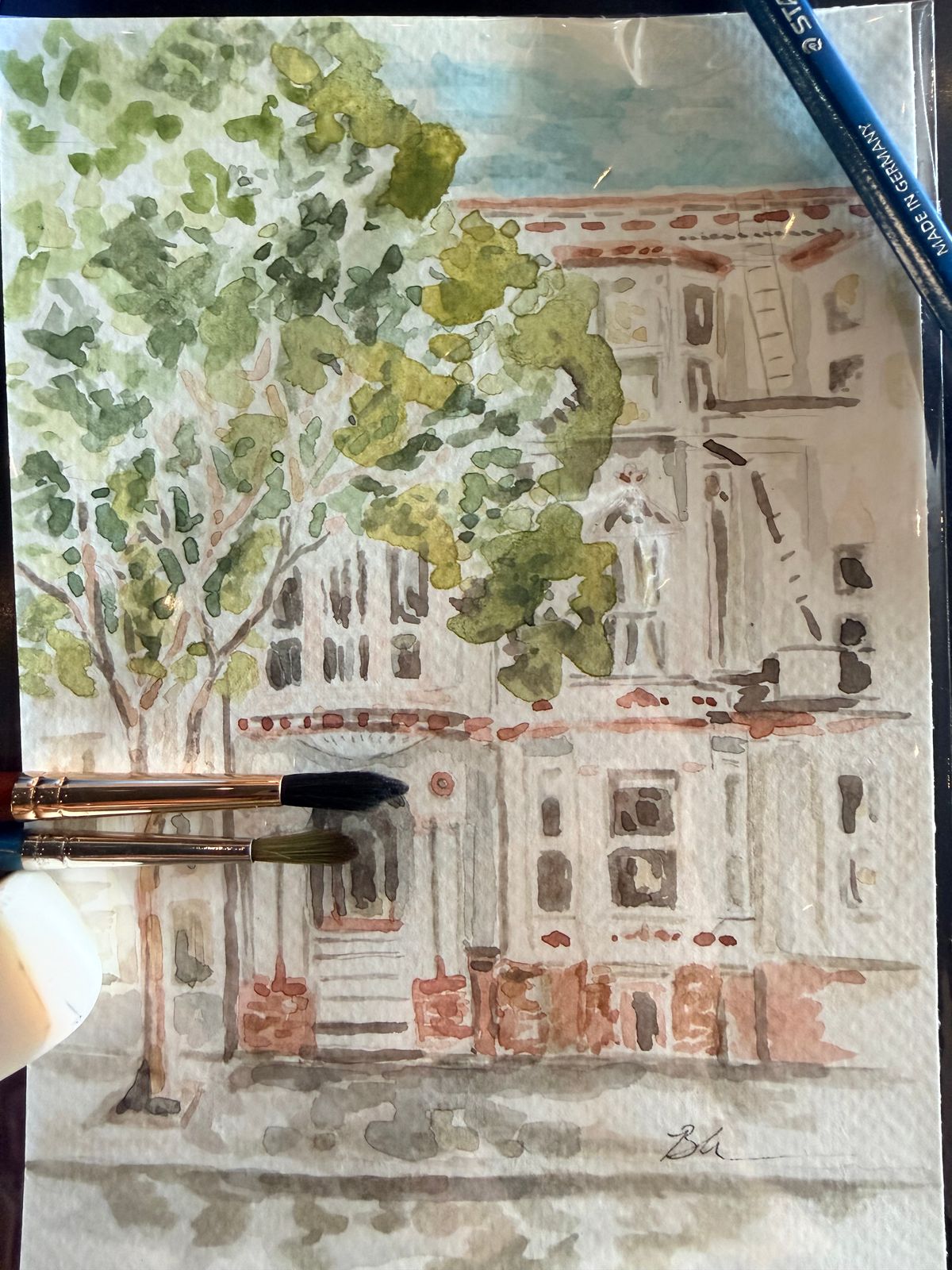 Watercolor of a pale multi-story building with reddish-brown trim beside a leafy green tree, shown on the worktable with two paintbrushes and a blue pencil resting on the paper